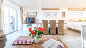 Un salón amplio con televisor, sillas de mimbre, una mesa de comedor con un jarrón de flores y una zona de cocina con encimera y armarios.