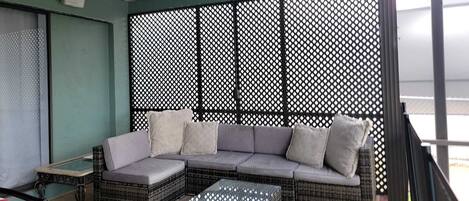 A balcony with a wicker sofa, cushions, and a glass coffee table.