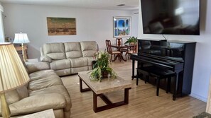Una sala de estar con sofá, mesa de centro con plantas, mesa de comedor con sillas, piano y televisor.