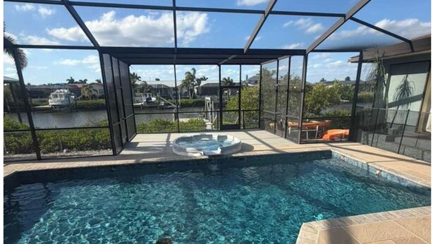 A swimming pool with a glass enclosure, clear blue water, and a view of a lake and buildings in the background. - Grand Oak Resort Waterfront Pool/Spa/2 King BR/kayak/Pet extended stay discounts (Apollo Beach)
