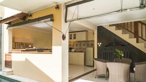 A poolside area with wicker furniture, a kitchen in the background, and a staircase leading upstairs. - Studio Bawa (Seminyak)