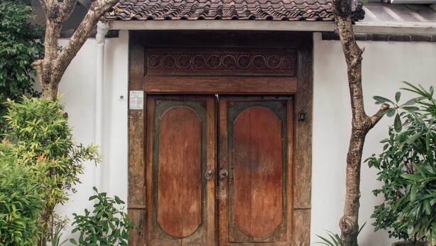 A traditional wooden door with a tiled roof above it, flanked by trees and potted plants. - Studio Bawa (Seminyak)