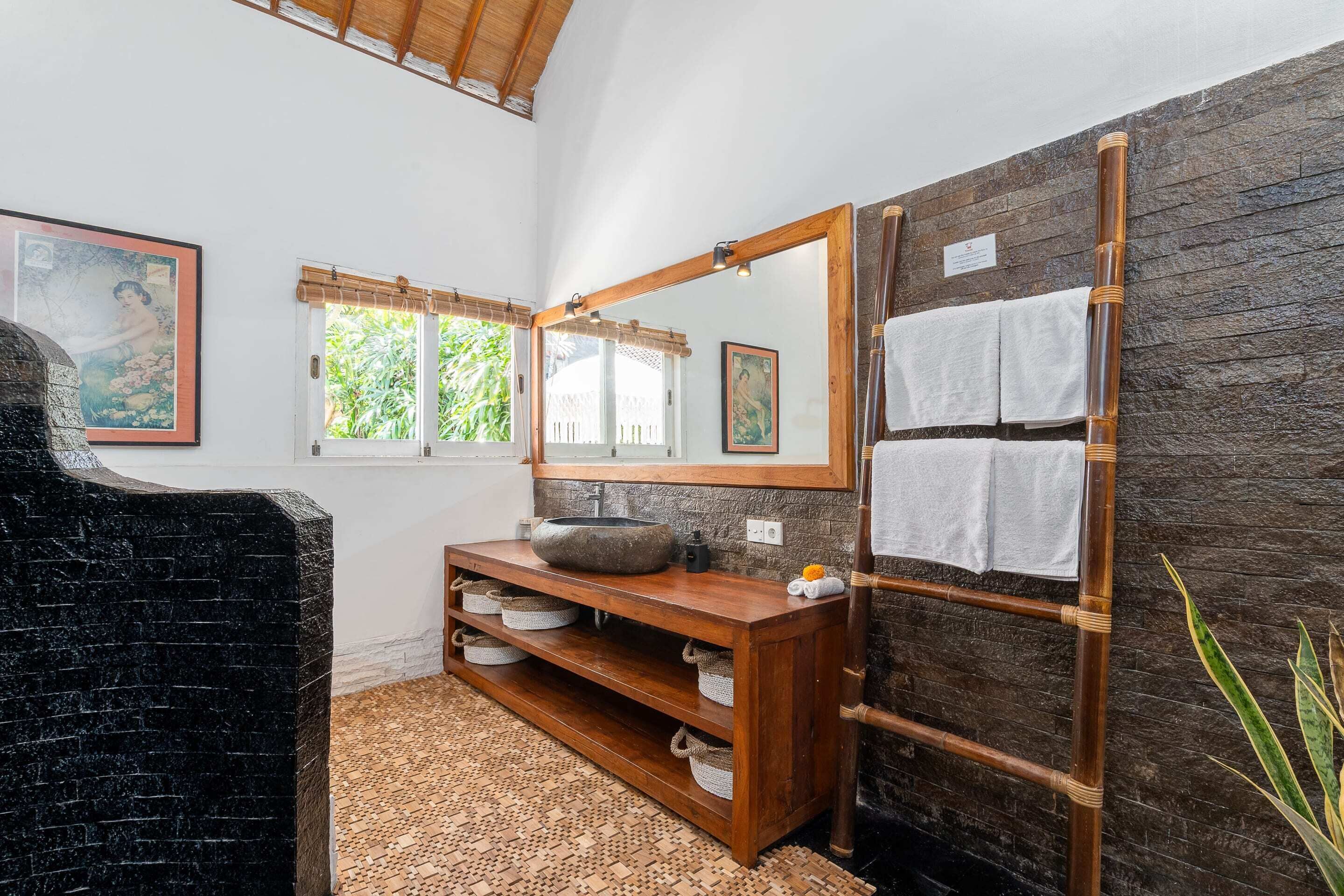 A bathroom with a stone sink, wooden vanity, and a large mirror. There are towels hanging on a bamboo rack and a framed picture on the wall.