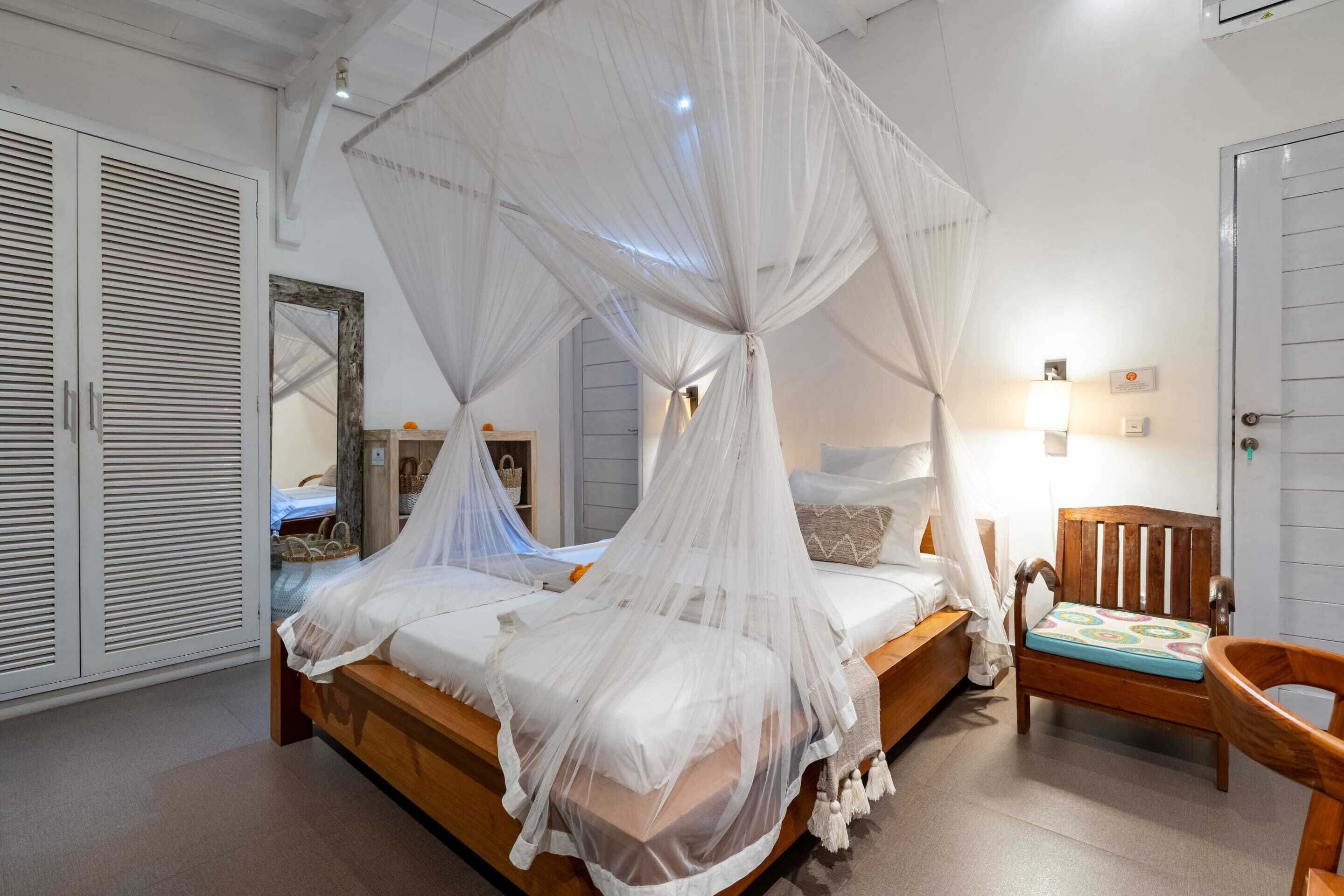 Un dormitorio con una cama cubierta por un dosel blanco, una silla de madera y una mesita de noche con una lámpara.