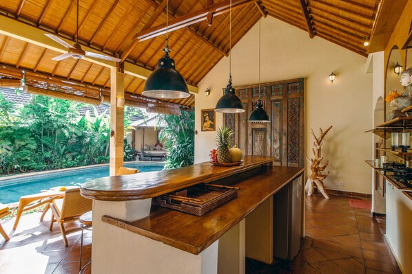 A kitchen with a wooden countertop, hanging pendant lights, and a view of a pool. - Villa Orchidea (Seminyak)