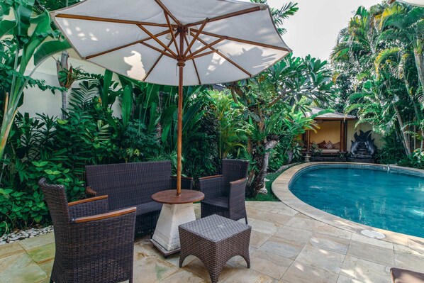 An outdoor seating area with wicker furniture and a large umbrella, surrounded by lush tropical plants and a pool. - Villa Orchidea (Seminyak)
