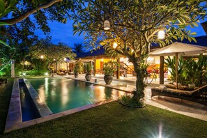 A pool area with a lounge chair and umbrella, surrounded by potted plants and a well-lit patio. - Villa Kiki (Seminyak)