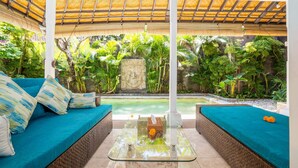 A covered outdoor seating area with a glass table, blue cushioned sofas, and a stone wall in the background. - Villa Shanti (Seminyak)