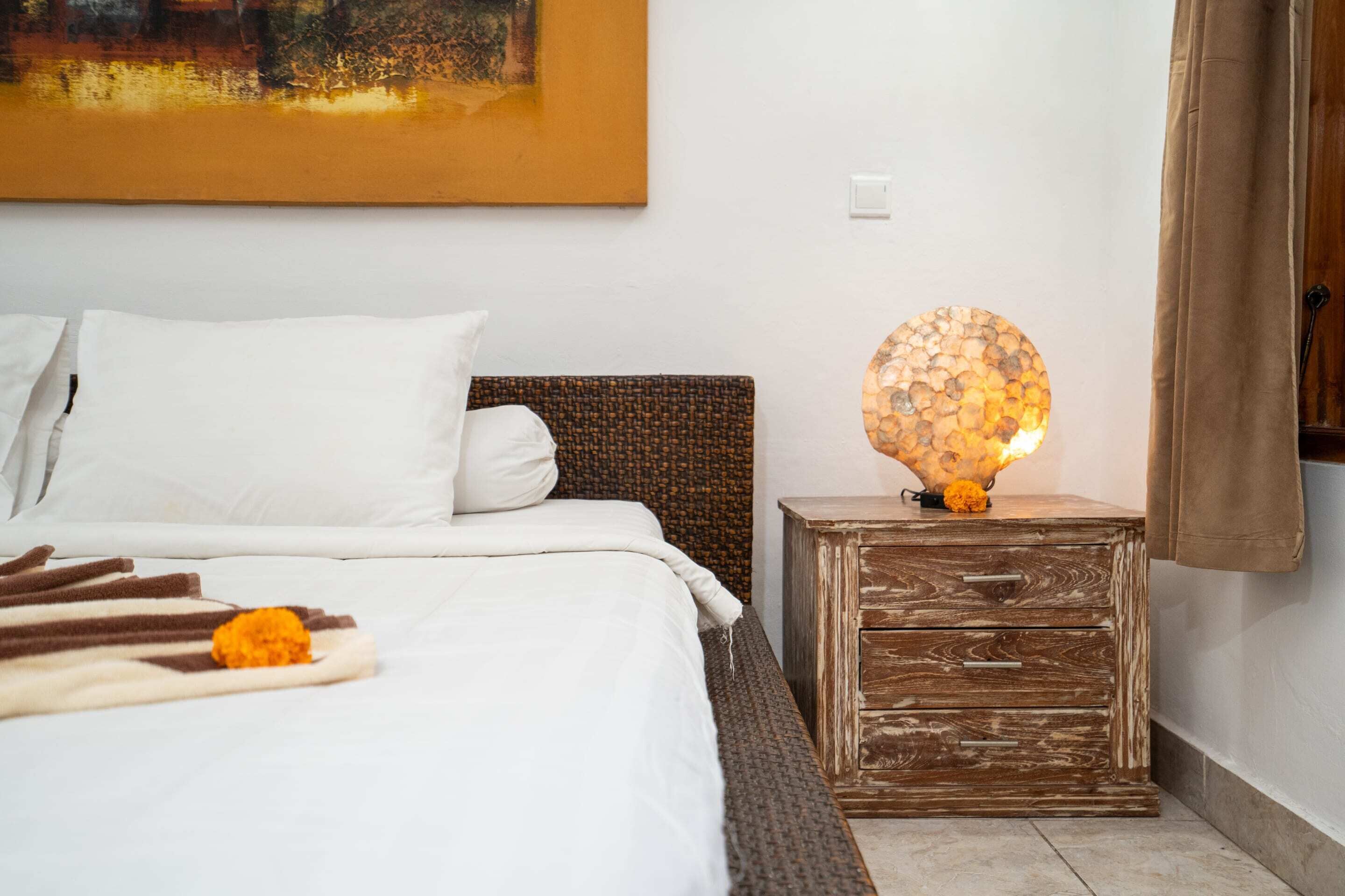 A neatly made bed with white linens and a decorative orange flower. There is a wooden nightstand with drawers and a textured lamp beside the bed.