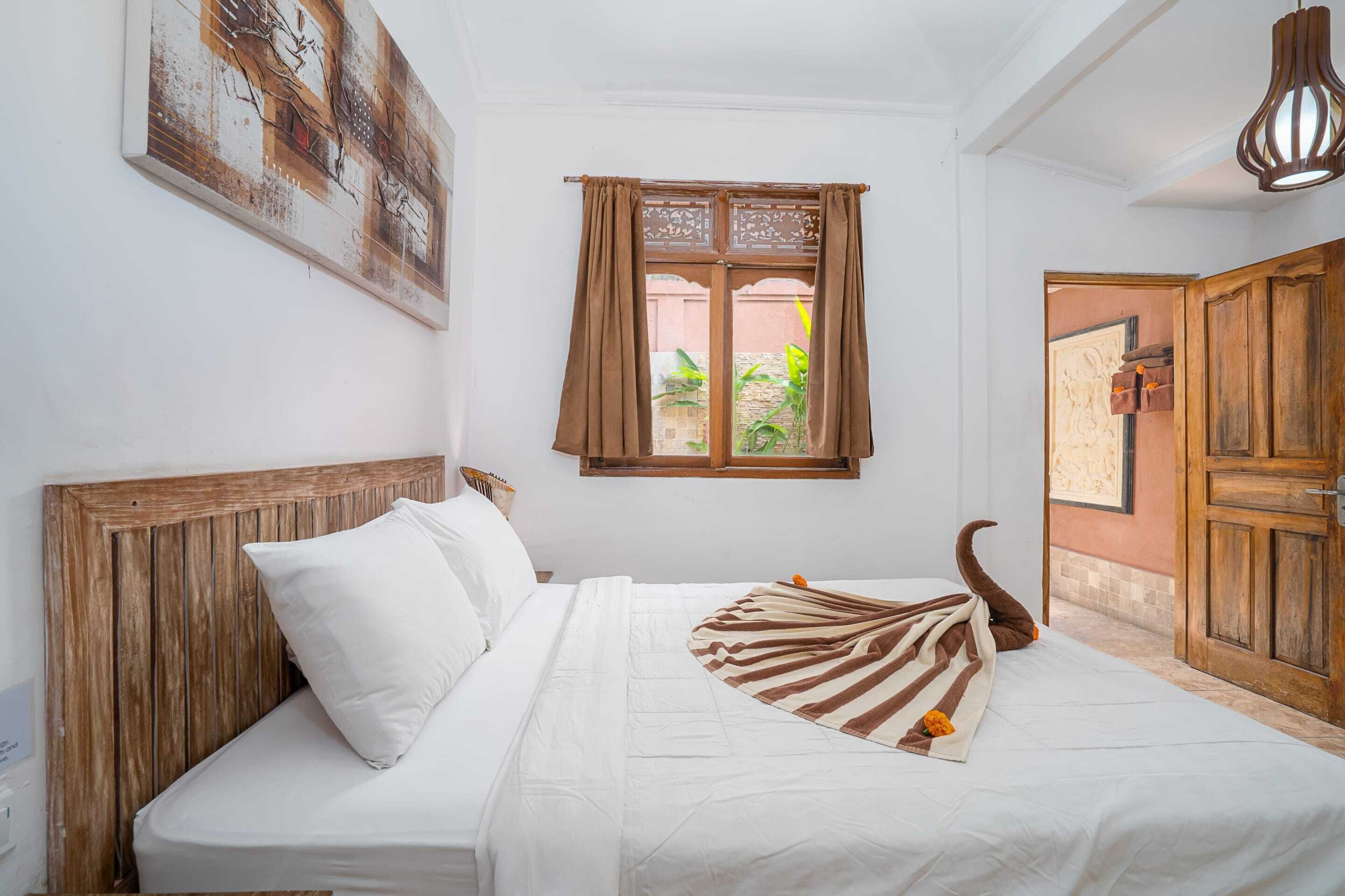 Een netjes opgemaakt bed met een gestreept dekbed en een decoratief kussen, een houten hoofdbord, een raam met gordijnen en een houten deur.