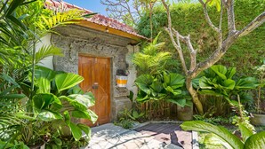 A stone building with a wooden door, surrounded by lush tropical plants and a tiled patio. - Villa Shanti (Seminyak)
