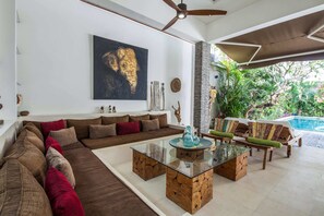 A modern living room with a large sofa, a glass coffee table, and a pool area visible through an open door.