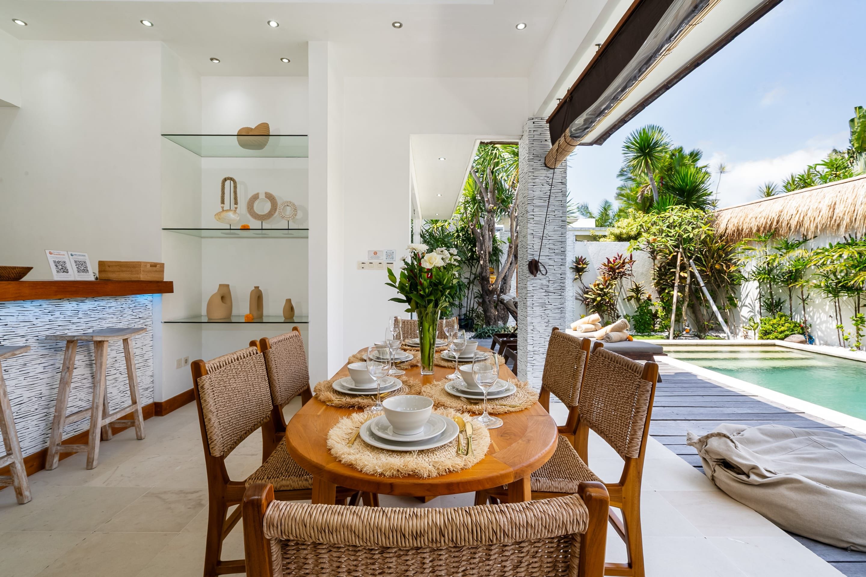 A dining area with a wooden table set for six, surrounded by wicker chairs, with a glass shelf displaying decorative items in the background.