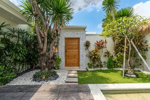 Une maison moderne avec une porte en bois, entourée de plantes tropicales et d’un petit jardin.