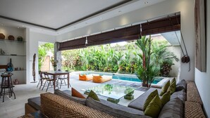 A modern living room with a pool view, featuring a sofa, coffee table, and dining area. - Villa Kambodja (Seminyak)