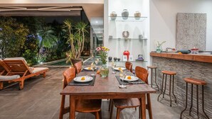 A dining area with a table set for four, surrounded by wooden chairs. There is a bar area with stools to the right. The room features a large glass window overlooking a garden with tropical plants. - Villa Laurent (Seminyak)