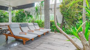 A wooden deck with four lounge chairs, a ceiling fan, and lush greenery in the background. - Villa Laurent (Seminyak)