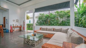 A modern living room with a glass coffee table, a sofa, and a view of lush greenery outside. - Villa Laurent (Seminyak)