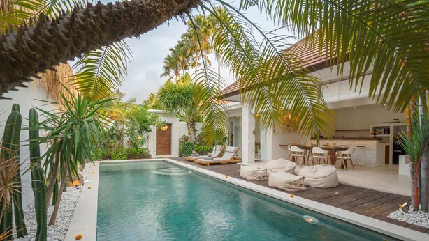 Un espace piscine doté de chaises longues et d’un coin repas, le tout entouré de plantes tropicales et d’un mur en pierre.