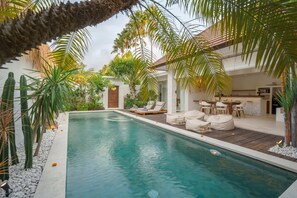 A pool area with lounge chairs and a dining area, surrounded by tropical plants and a stone wall. - Villa Astaga (Seminyak)