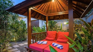 A wooden gazebo with a red cushioned bench and a small table, surrounded by lush greenery. - Villa Suliac (Legian)