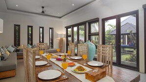 A dining area with a long wooden table set for a meal, surrounded by chairs, and large windows offering a view of the outdoors. - Villa Athena (Seminyak)