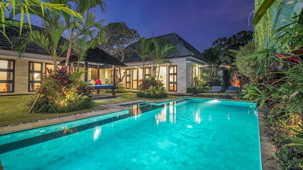 A swimming pool with a tiled deck, surrounded by lush greenery and a well-lit house with large windows. - Villa Athena (Seminyak)