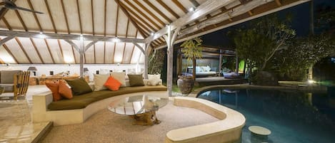 A poolside lounge area with a curved seating arrangement, a glass coffee table, and a view of the pool and surrounding greenery.