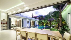 A modern outdoor dining area with a pool, surrounded by lush greenery and a view of neighboring houses. - Villa Jago (Seminyak)