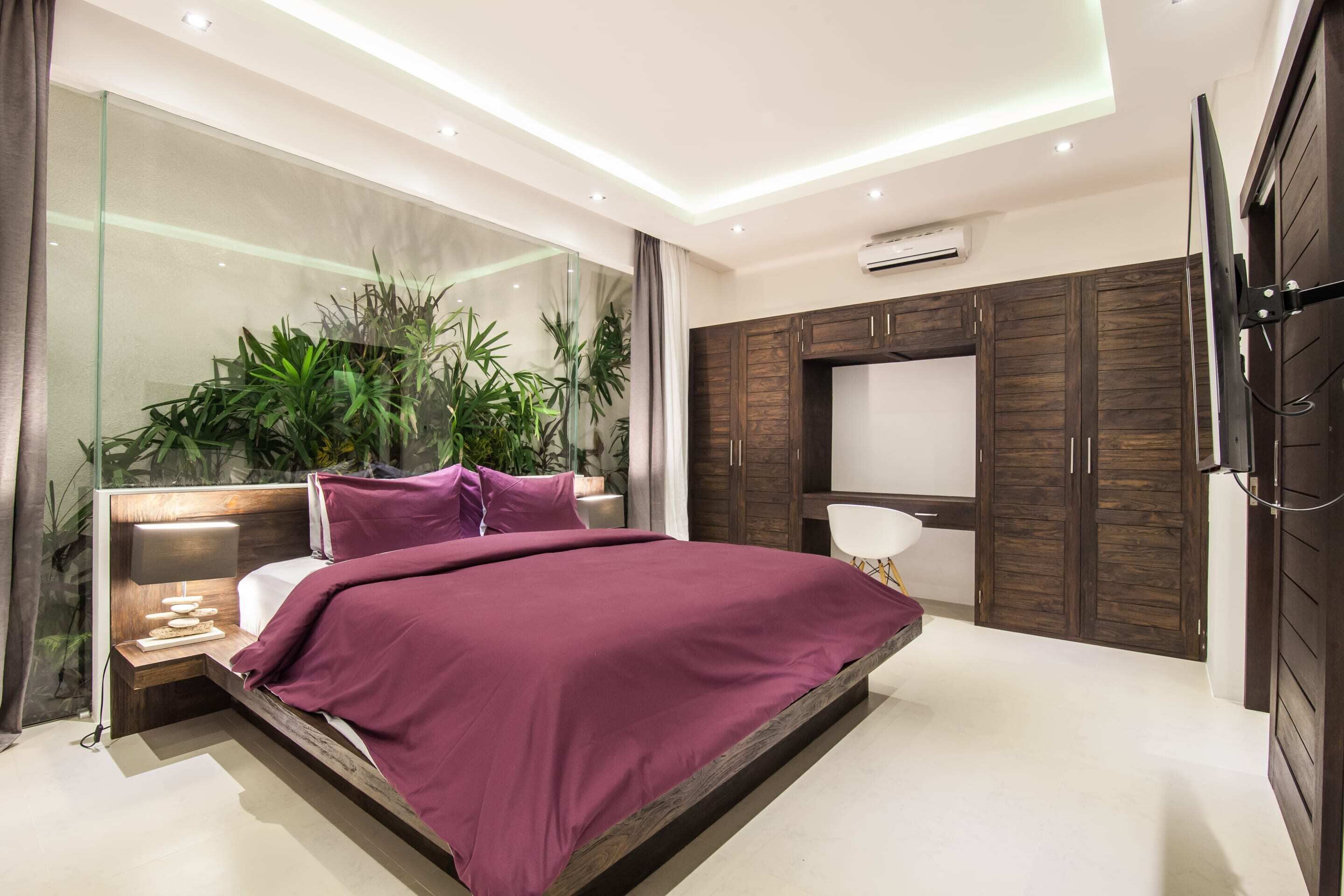 A modern bedroom with a glass wall, a bed with a purple bedspread, a bedside table with a lamp, and a TV mounted on a wooden cabinet.