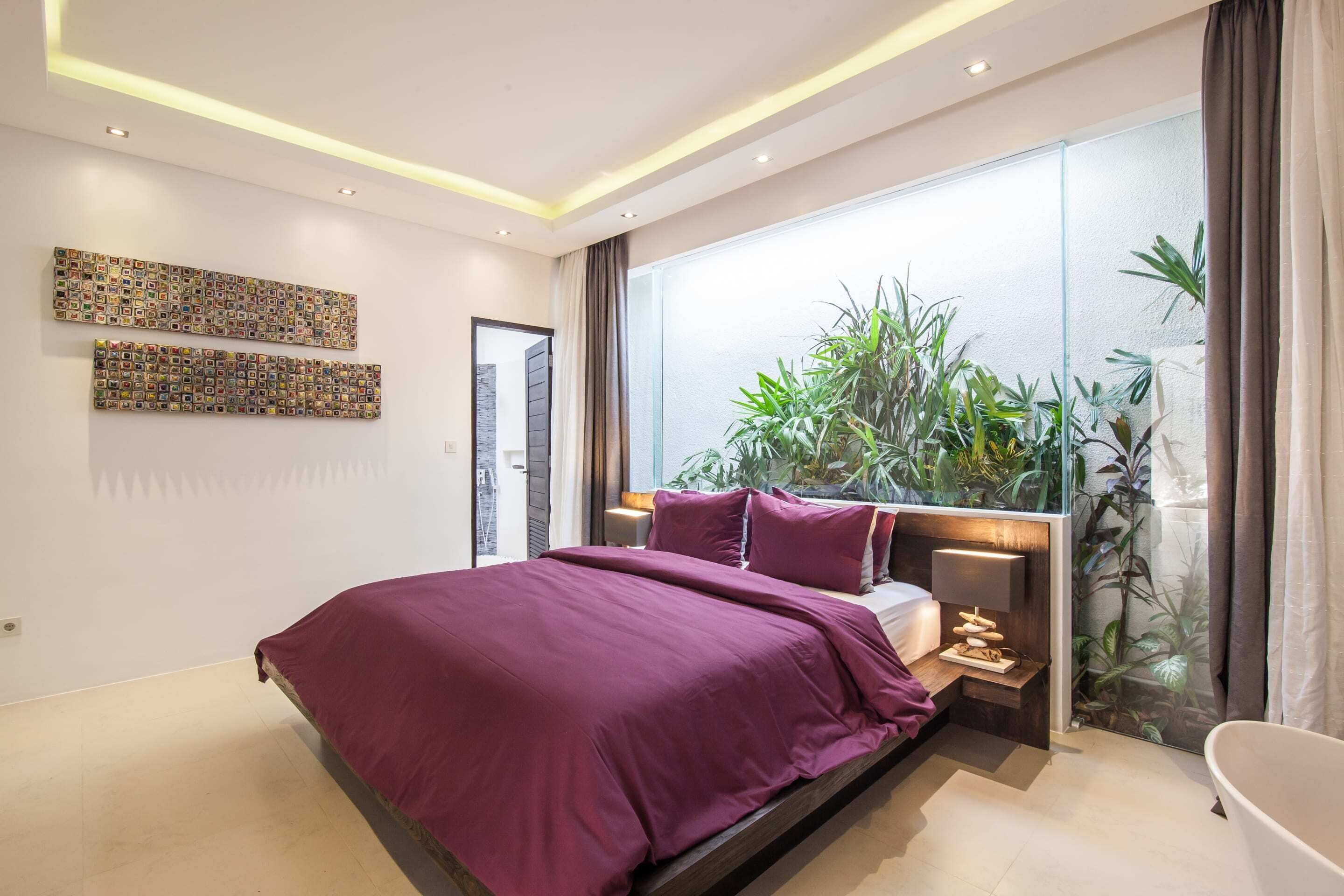 A modern bedroom with a large bed, a nightstand, a lamp, and a view of greenery through a sliding door.