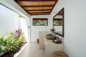 A bathroom with a stone sink, a mirror, and a window with a view of greenery. - Villa Bali Sari (Canggu)