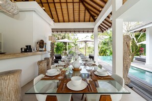 A dining area with a glass table set for a meal, surrounded by chairs, with a pool visible in the background. - Villa Bali Sari (Canggu)