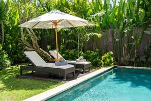 Zona de piscina con sillas de descanso y una sombrilla, rodeada de abundante vegetación.