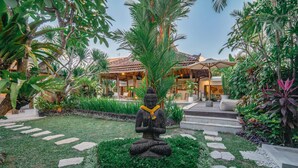 A statue of a meditating figure surrounded by a garden with a pathway leading to a building with a tiled roof. - Villa Marula (Seminyak)