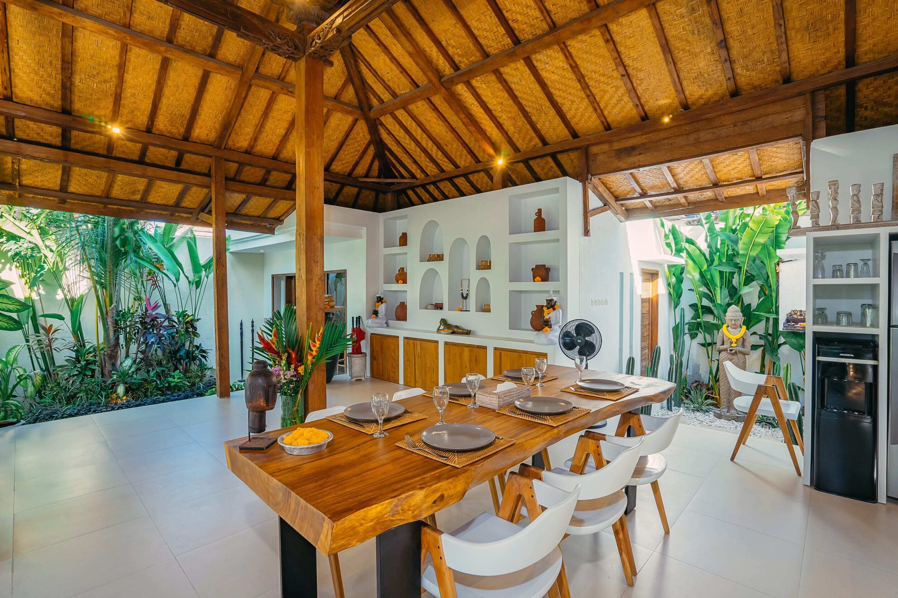 A spacious dining area with a wooden table set for six, surrounded by white chairs. A kitchen with white cabinets and a refrigerator is visible in the background.