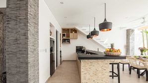 A modern kitchen with a central island, black pendant lights, and a dining area with wicker chairs. - Villa Clemence (Canggu)