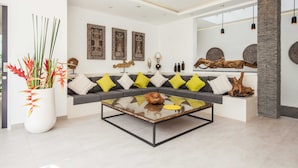 A modern living room with a glass coffee table, grey sofa with yellow cushions, decorative wall art, and a large potted plant. - Villa Clemence (Canggu)