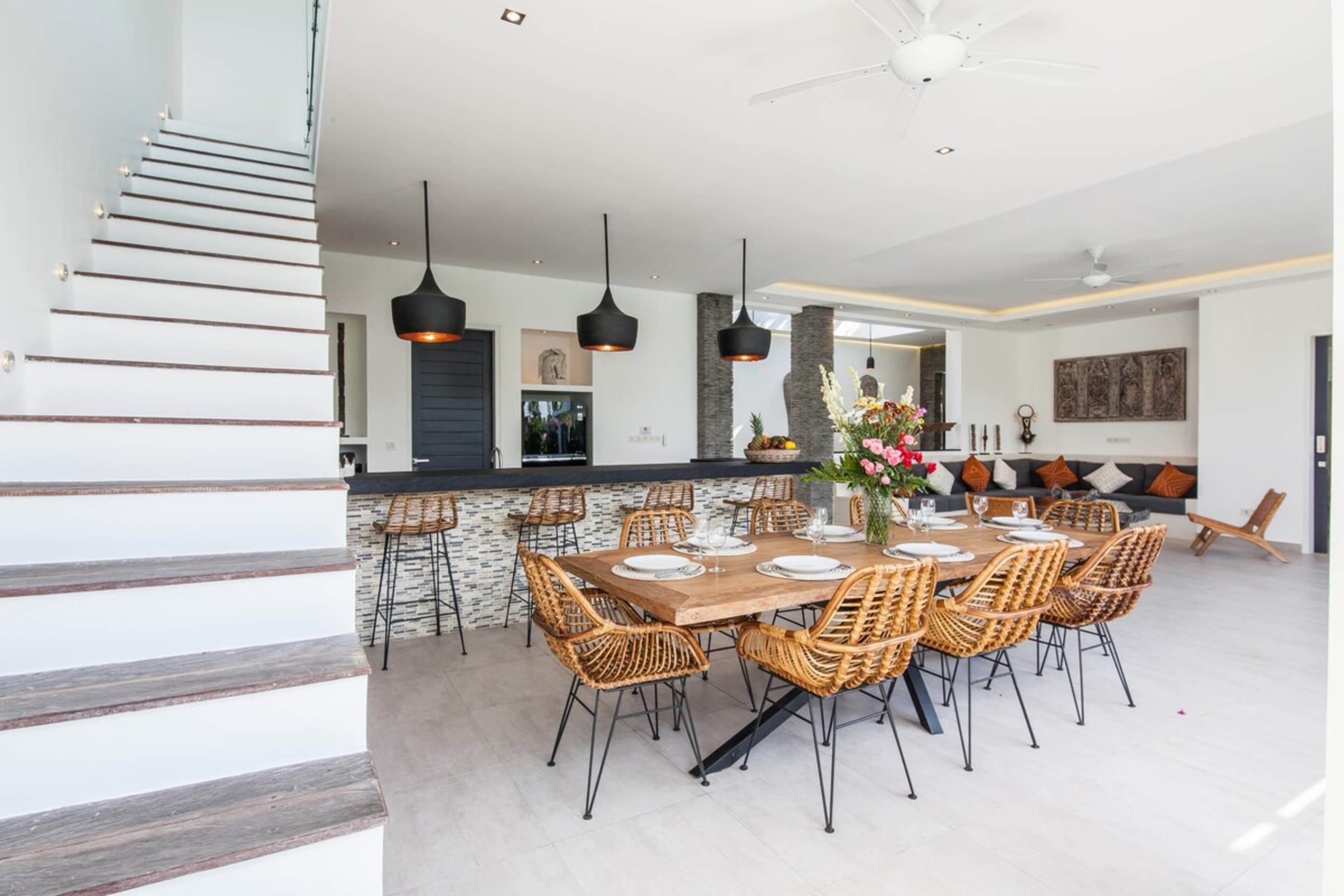 A spacious dining area with a large wooden table and wicker chairs, a kitchen with a stone backsplash, and a modern staircase.