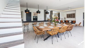 A spacious dining area with a large wooden table and wicker chairs, a kitchen with a stone backsplash, and a modern staircase. - Villa Elisa (Canggu)