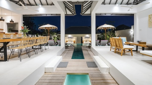 A modern outdoor dining area with wooden tables and chairs, a central pathway, and umbrellas. - Villa Ananda (Canggu)