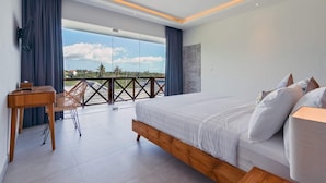 A modern bedroom with a large bed, a wooden desk, and a balcony with a view of the sky and trees. - Villa Ananda (Canggu)