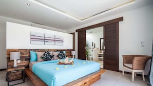 Un dormitorio moderno con cabecera de madera, una cama grande con colcha azul, una mesita de noche con lámpara y una silla de mimbre.