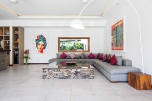 A modern living room with a stone coffee table, grey sofa with colorful cushions, a large window, and a painting on the wall. - Villa Gala (Seminyak)