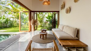 Un salon moderne avec un canapé, une table basse ronde et une vue sur un jardin luxuriant et une piscine.