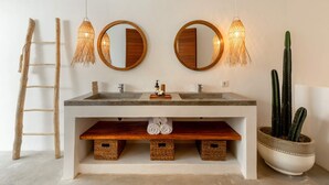 Une salle de bain avec deux lavabos, une échelle en bois, des luminaires en fibres tressées et un cactus en pot.