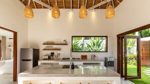 A modern kitchen with a central island, wicker chairs, and a large window overlooking a garden. - Villa Lumahi Tiga (Cemagi)