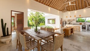 A spacious living area with a wooden dining table and chairs, a kitchen with modern appliances, and a large window offering a view of the outdoors. - Villa Lumahi Tiga (Cemagi)