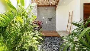 A modern outdoor shower area with a concrete wall, wooden deck, and surrounding lush greenery. - Villa Lumahi Tiga (Cemagi)