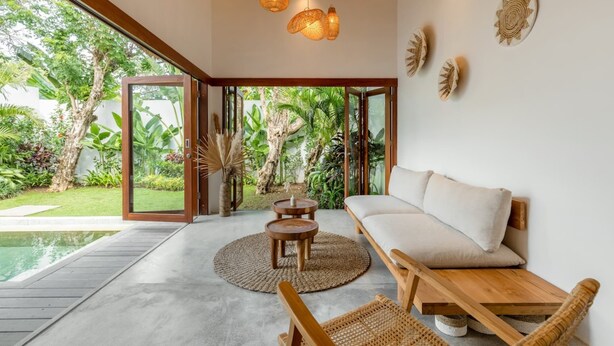 A modern living room with a large glass door leading to a lush garden, a wooden bench, a round wooden table, and a sofa with cushions. - Villa Lumahi Dua (Cemagi)